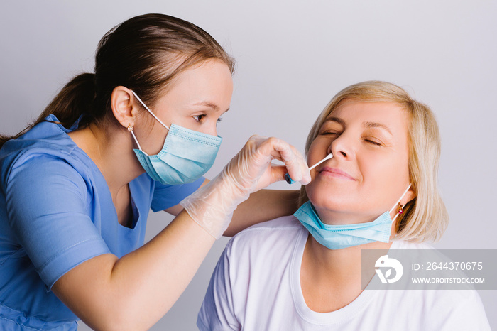 A young doctor in a medical mask takes a COVID 19 test, nasal swab from an elderly woman against a white background. Virus detection concept.