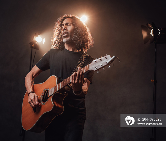 Middle aged hispanic musician in black t-shirt emotionally singing and playing guitar. View of musician in the spotlight