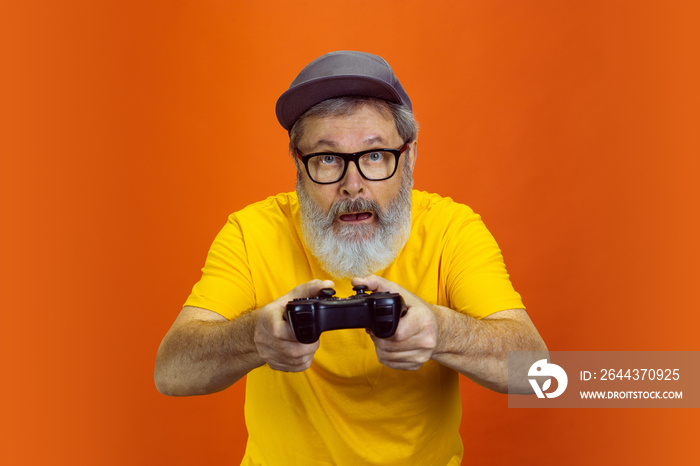 Gamer man. Portrait of senior hipster man using devices, gadgets isolated on orange studio background. Tech and joyful elderly lifestyle concept. Trendy colors, forever youth. Copyspace for your ad.