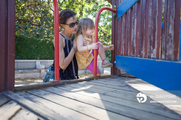 mother woman helping three years old blonde girl, with yellow dress and pink tights, to climb on wooden playground, in park of Madrid city