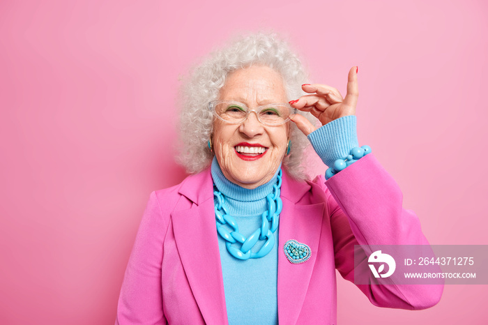 Portrait of smiling good looking senior lady keeps hand on rim of spectacles wears fashionable clothes happy to hear something pleasant has glad expression. Age old people fashion and style.