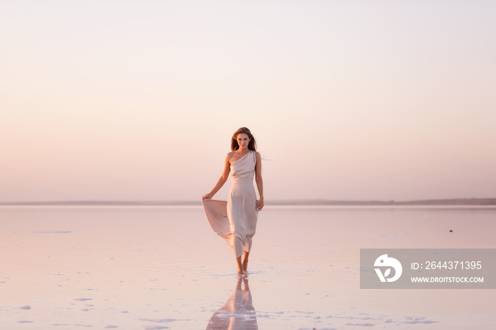 Young blonde woman in an evening airy pastel pink, powdery dress stands barefoot on white crystallized salt. Girl with natural make-up, hair is developing. Salt mining trip, walking on water at sunset