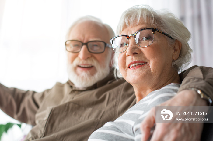 Portrait of mature lady and old happy man hugging. Focus on woman while they are laughing sitting on couch at home