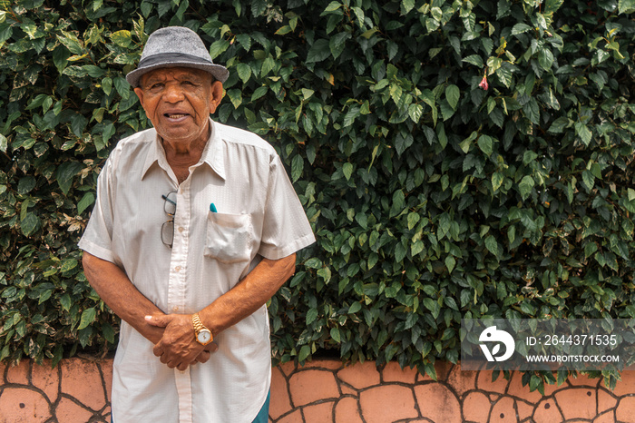 Nicaraguan old man in a hat and short-sleeved shirt looking at the camera with plants in the background