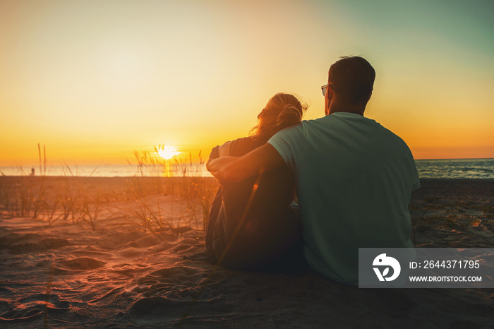 young lovers couple sitting in sand on beach at romantic golden sunset. back view