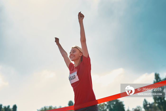 Low angle view of excited young female athlete with arms raised reaching the finish line at track field during marathon outdoors