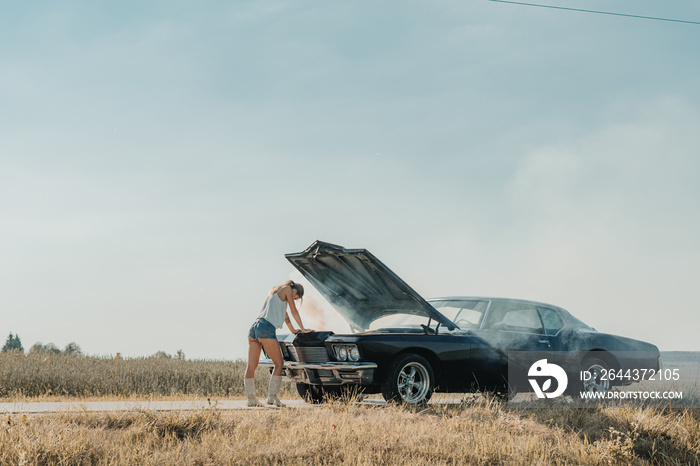 Young female standing near overheated car in the field, bright sunlight, steam under the hood