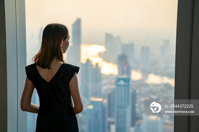 Asian woman standing by the window at skyscraper office building in metropolis and looking cityscape at summer sunset. Beautiful female relax and enjoy outdoor lifestyle activity in the city at night