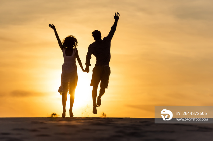 silhouettes of man and woman jumping on beach against sun during sunset