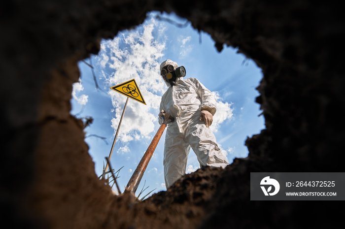 Below view on male ecologist digging pit by shovel, wearing protective overalls and gas respirator near biohazard symbol warning about dangerous biological materials. View from inside pit.