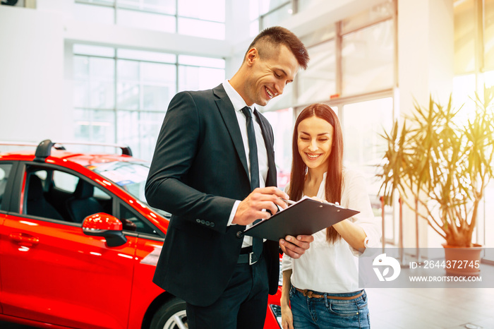 Sign here please. The dealer manager in a black suit gives documents for buying a car to a young smiling woman