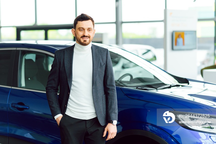 Handsome bearded buyer in casual wear in dealership, guy looks on camera while standing near car with crosed arms