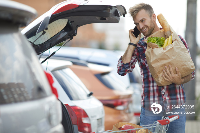 Portrait of handsome young man speaking by phone while packing groceries into car trunk , copy space