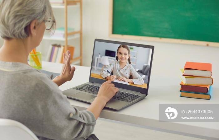 Home schooling. Mature teacher talking to little girl on laptop from classroom. Tutor communicating with pupil online