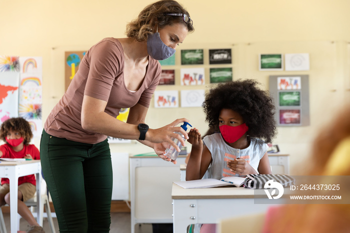 Female teacher wearing face mask sanitizing hands of girl in class