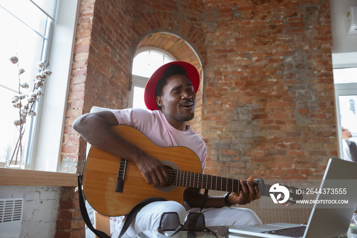 African-american musician playing guitar during online concert at home isolated and quarantined. Using camera, laptop, streaming, recording courses. Concept of art, support, music, hobby, education.