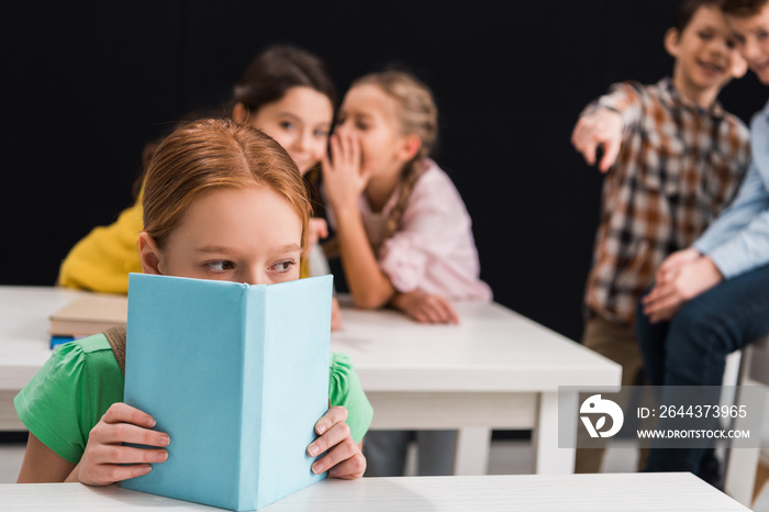 selective focus of upset schoolkid covering face with book near classmates gossiping and pointing with finger isolated on black, bullying concept