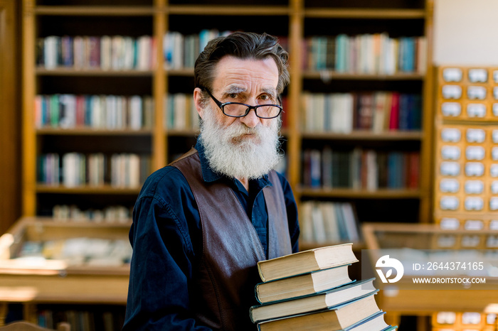 Portrait of handsome senior bearded retired man, librarian or teacher, choosing books in library, holding stack of books, looking at camera, book shelves on the background