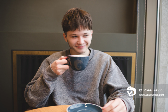 Smiling teen boy generation Z 16-17 year old drinking fresh tasty coffee sitting in cafe indoors. Handsome blond teenage hipster.