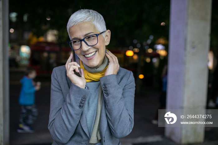 Smiling mature senior woman with short gray hair and eyeglasses use phone on street, night scene in city