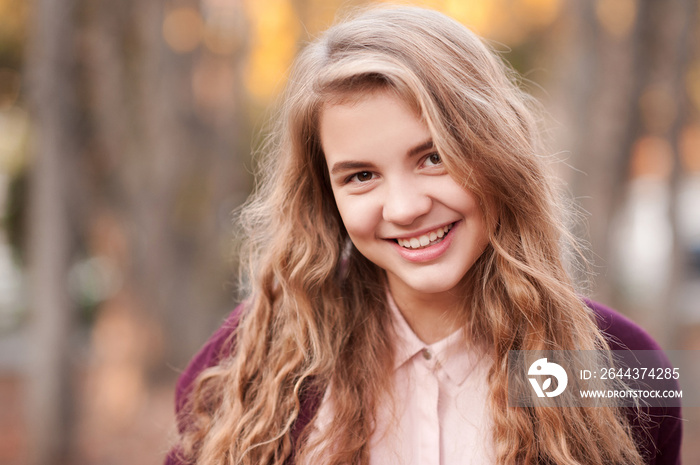 Smiling blonde teenage girl 14-16 year old with curly long hair outdoors. Looking at camera. Beautiful portrait. Happiness.