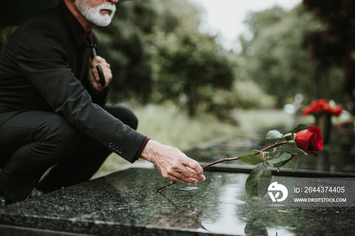 Elegant sad elderly man standing on the rain with umbrella and grieves at the grave of a loved person