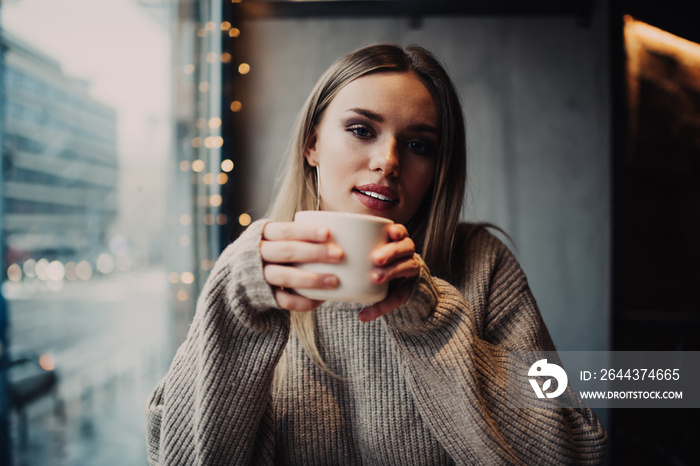 Portrait of attractive female customer enjoying weekend time for visiting coffee shop with aroma cappuccino for warming, beautiful Caucasian hipster girl 20 years old with tea cup looking at camera