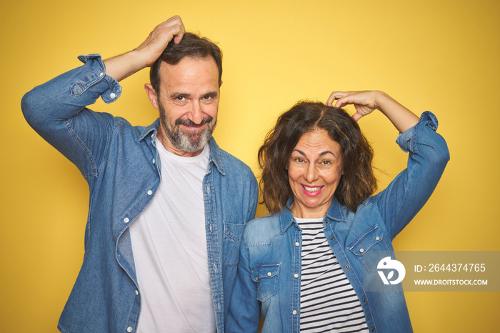 Beautiful middle age couple together wearing denim shirt over isolated yellow background confuse and wonder about question. Uncertain with doubt, thinking with hand on head. Pensive concept.