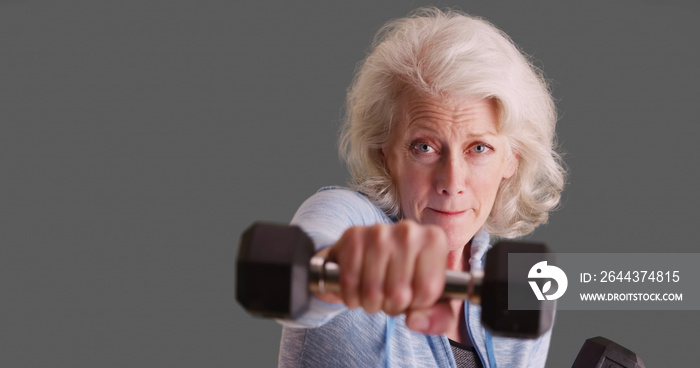 Determined senior woman exercising with dumbbells looking at camera on gray backdrop