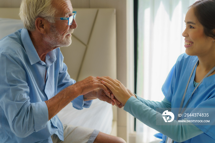 Asian nurse standing on a home bed next to an older man helping hands, care. Elderly patient care and health lifestyle, medical concept.