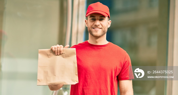 Young caucasian deliveryman smiling happy holding delivery paper bag at the city.