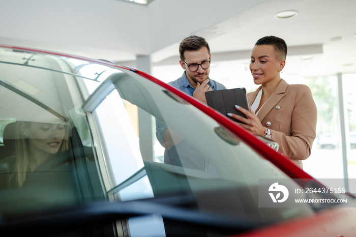 Young smiling saleswoman is holding a tablet and showing new car to couple in showroom.