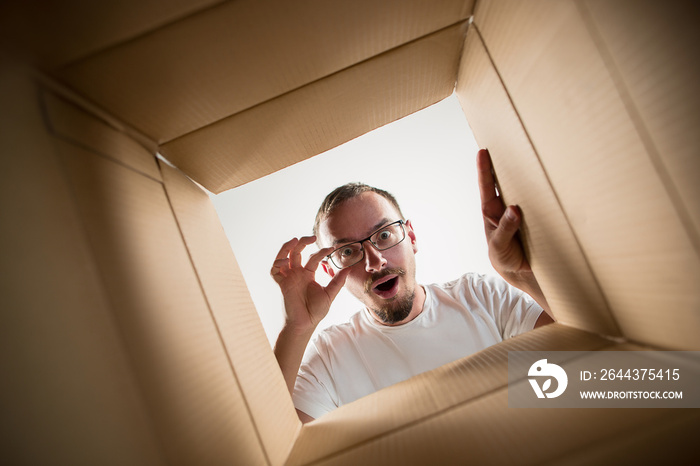 The surprised man unpacking, opening carton box and looking inside. The package, delivery, surprise, gift lifestyle concept. Human emotions and facial expressions concepts