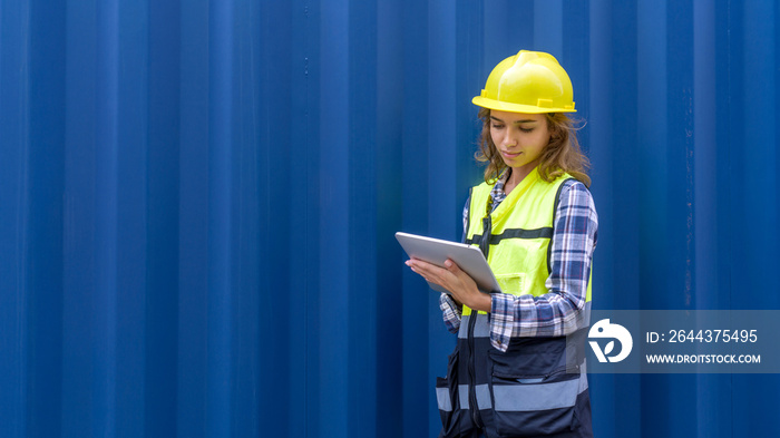 Young caucasian woman with safety vest and yellow hardhat checking shipping schedule on tablet computer, planning for next shipment. A large cargo container is in the background