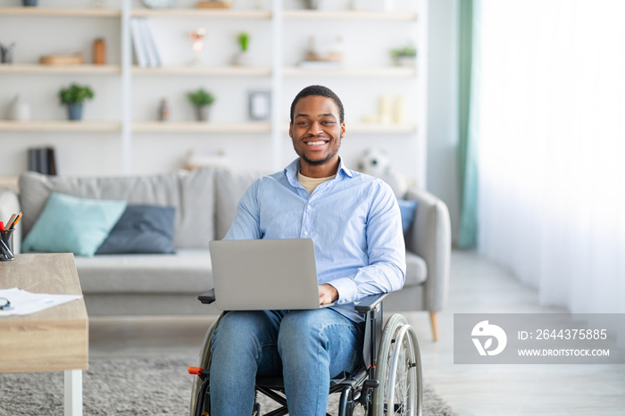 Portrait of joyful impaired young man in wheelchair using laptop for remote work at home