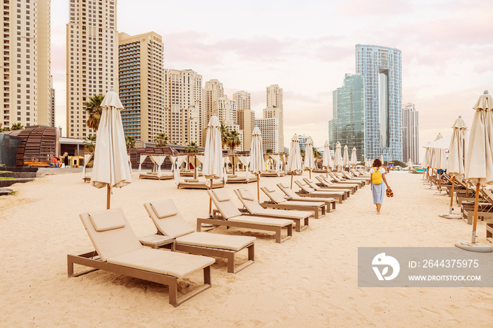 Woman tourist walks barefoot in the sand between rows of sun loungers and beach umbrellas in the JBR district in Dubai. Holidays and vacations in the UAE