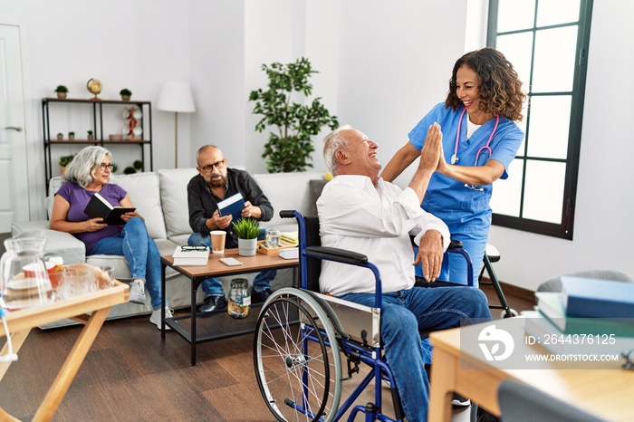 Middle age doctor woman and retired man sitting on wheelchair high five at nursing home.