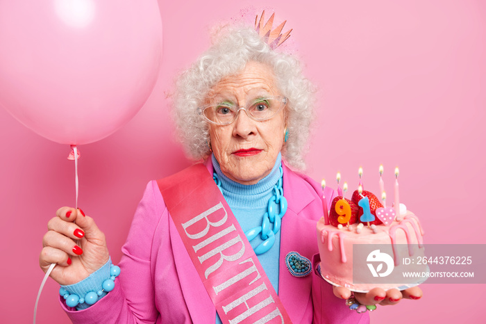 Portrait of serious ninety one years old woman celebrates birthday in family circle holds cake with candles and inflated balloon wears crown on head and festive clothes accepts congratulations