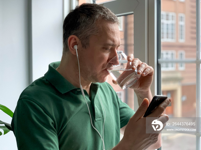 Middle aged man is talking on phone with friends or family, video call, watching video or checking news on social media. Staying at home by the window, wearing green polo t-shirt. Hangover, pure life