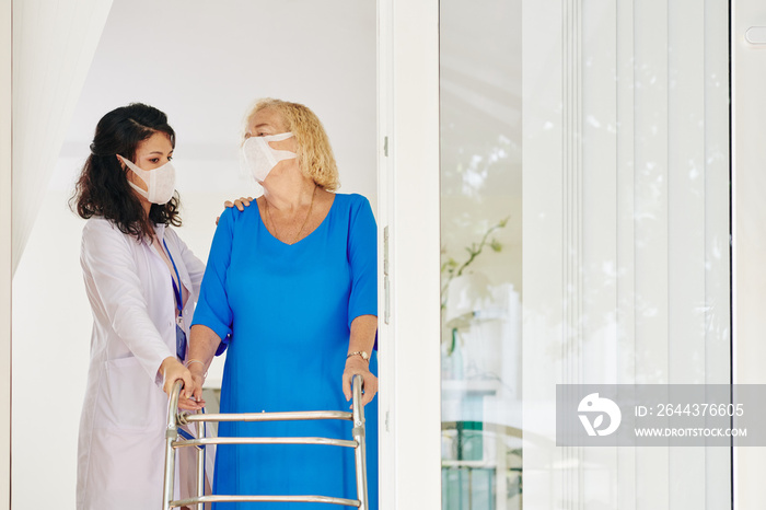 Serious young medical worker supporting senior patient moving around with walker