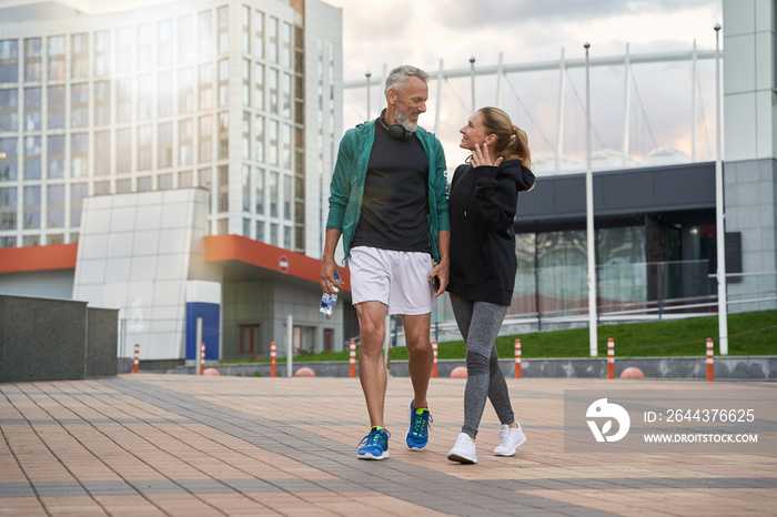 Full length shot of happy sportive middle aged couple in sportswear looking cheerful while walking together outdoors after workout