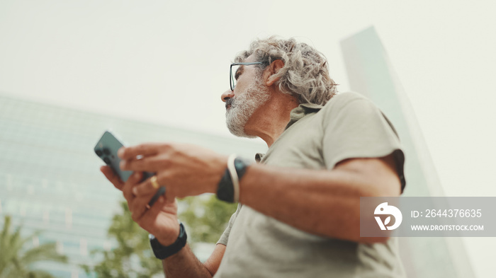 Friendly middle aged man with gray hair and beard looking at map trying to find his way using his mobile phone. Mature gentleman in eyeglasses using map app in cellphone outdoors