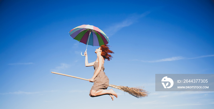 Young redhead witch on broom flying in the sky with umbrella