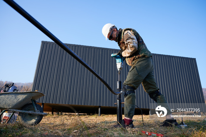 Male worker building pile foundation for wooden frame house. Man builder in white safety helmet drilling piles into the ground on blue sky background.