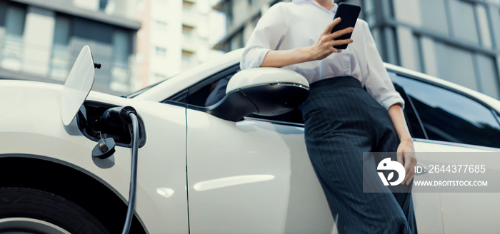 Closeup progressive businesswoman with smartphone, leaning electric car and charging station before driving around city center. Eco friendly rechargeable EV car powered by sustainable and clean energy