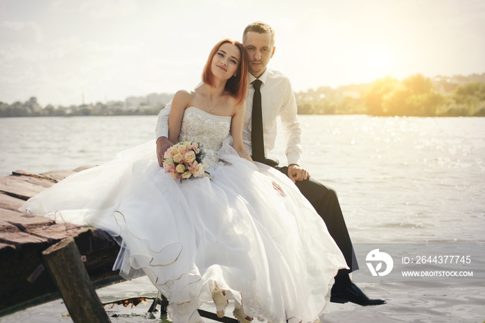 Wedding couple sitting on bridge near lake on sunset at wedding day. Bride and groom in love