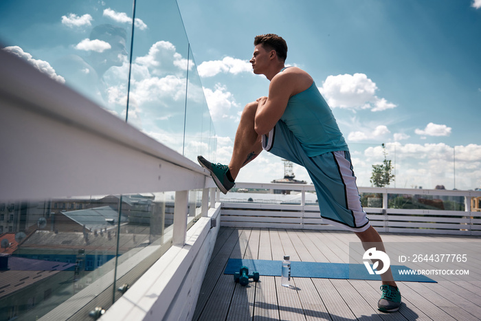 Athlete is putting foot on roof railing and staying in lunge for warming-up. He is preparing for training with dumbbells on top of house in city. He is looking far away while feeling concentrated