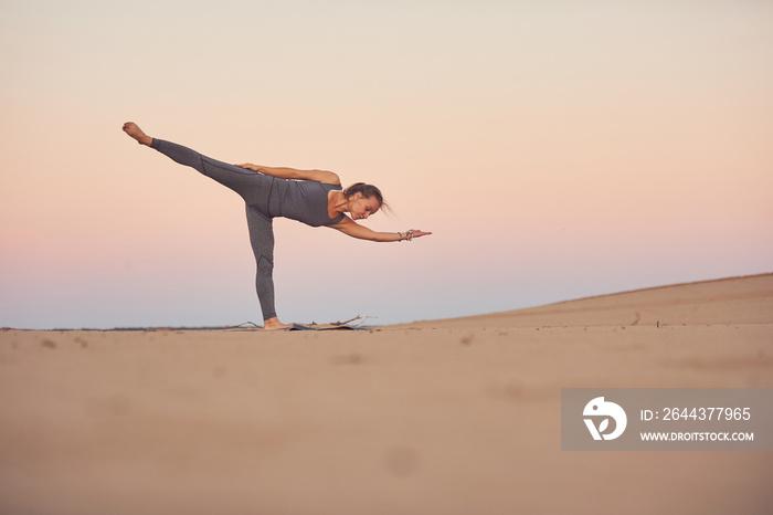 Beautiful young woman practices yoga asana Ardha Chandrasana - Half Moon pose in the desert at sunset