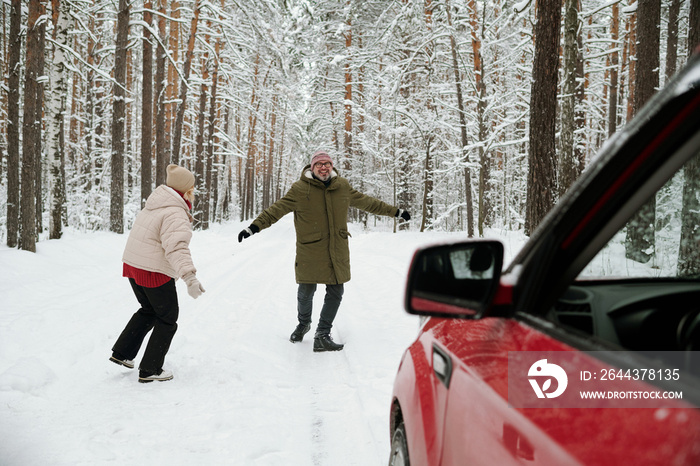 Playful mature couple in winterwear running along road in pinetree forest not far from their car while enjoying winter vacation or weekend