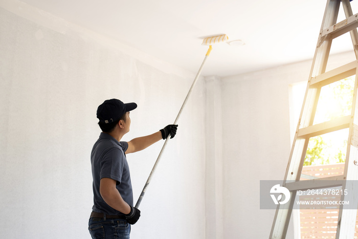 Asian rear view of a male painter drawing a wall with paint roller and a separate tank from a large empty space with wooden stairs.